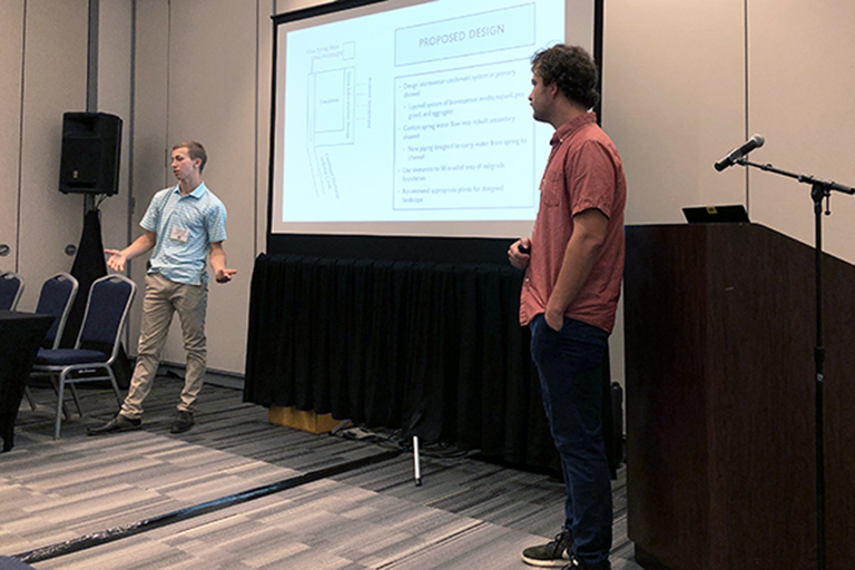 Charlie Cianciolo and Matthew Howard stand in front of a crowd, presenting at WEFTEC.