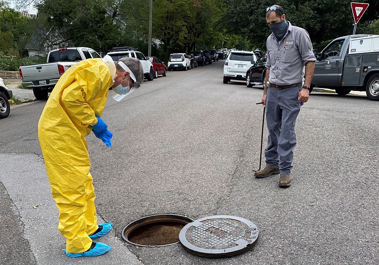 Two people pull samples from a wastewater access point in a street.