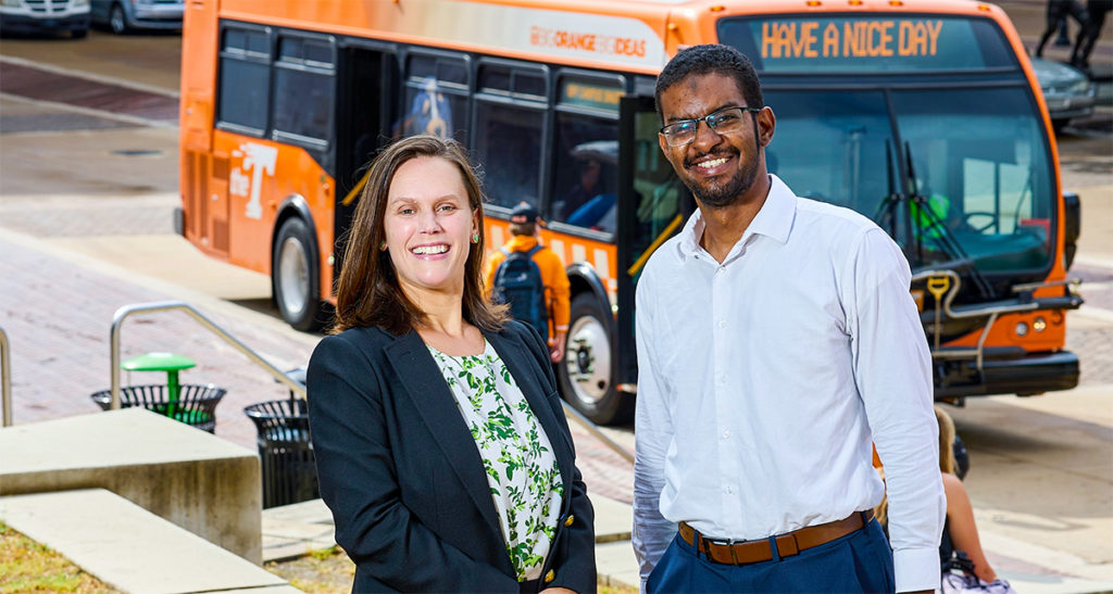 Candace Brakewood and a graduate student standing to the side of an orange bus driving on the road.