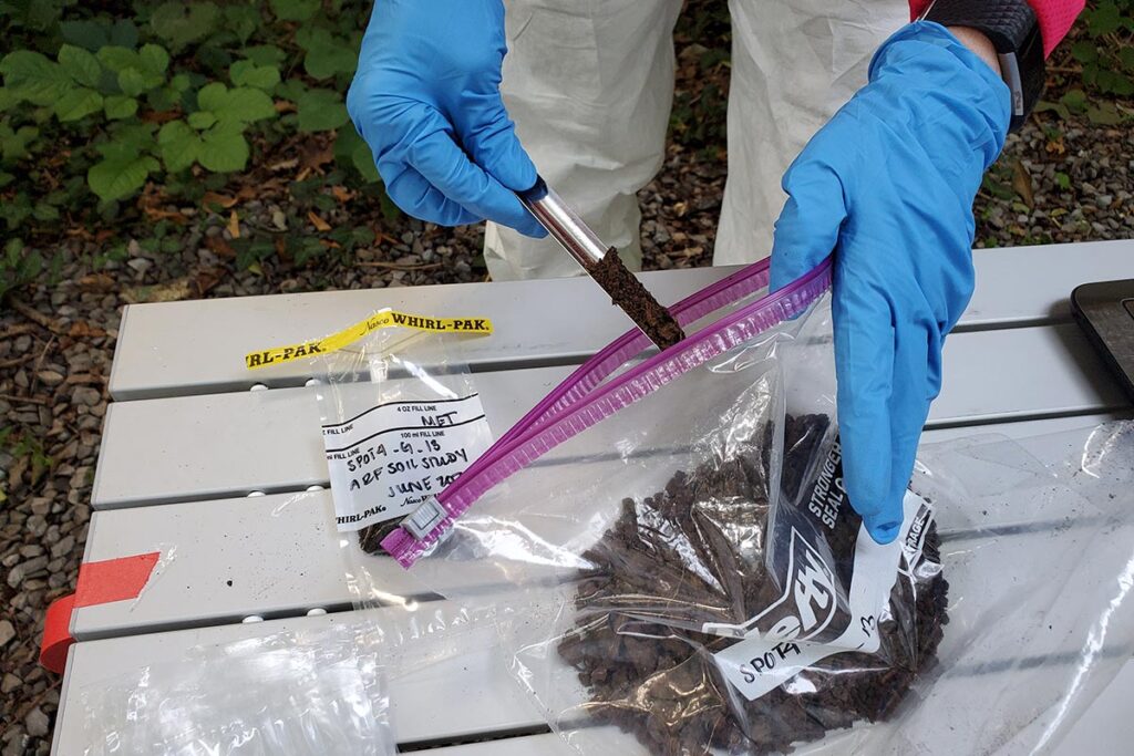 A researchers gloved hands use a tool to scoop soil from a larger sample contained within a plastic Ziploc bag which is resting on a white slat table in an outdoor lab environment at the Anthropological Research Facility.
